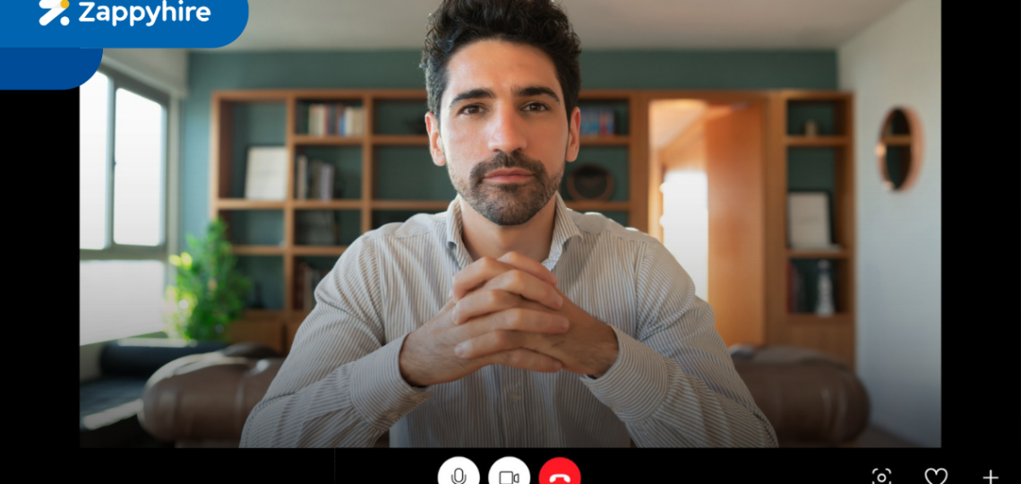 Screenshot of a one-way video interview interface with a professional man sitting in a well-lit, organized room, ready for the interview. The Zappyhire logo is visible at the top left corner of the interface, with controls for microphone, video, and call at the bottom of the screen.